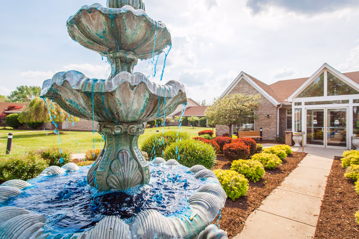Tiered stone fountain in the foreground with a landscaped walkway leading to the brick entrance of a residential care building.