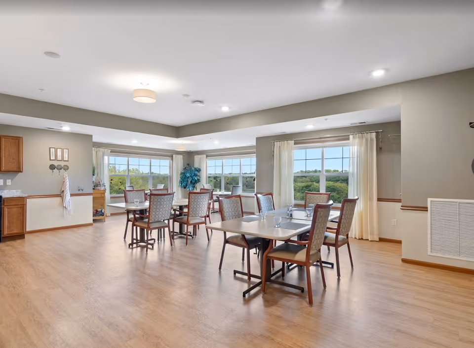 A bright dining room with wooden floors, several tables and chairs arranged neatly, large windows with sheer curtains allowing natural light and a view of greenery outside. The room has neutral-colored walls and ceiling lights.