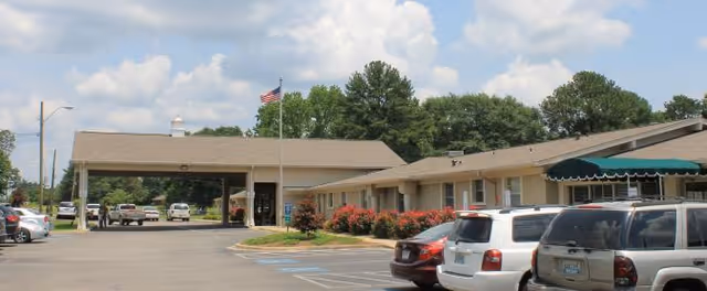 Exterior view of Ridgeview Health Services building with a covered entrance, an American flag on a flagpole, several parked cars in the parking lot, and trees in the background under a partly cloudy sky.