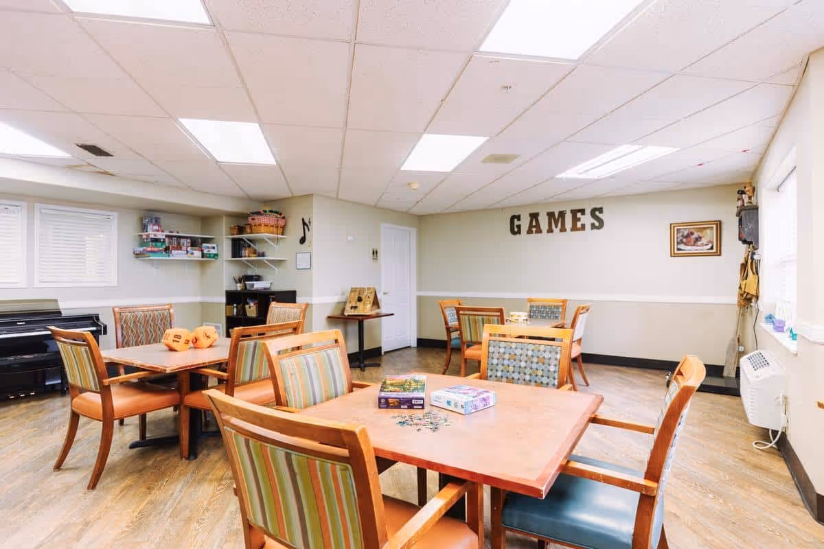 A bright game room in a senior living facility with several wooden tables and chairs arranged for playing board games and puzzles. The walls are light-colored with the word 'GAMES' displayed prominently. Shelves hold board games and other game supplies, and a piano is visible in the corner.