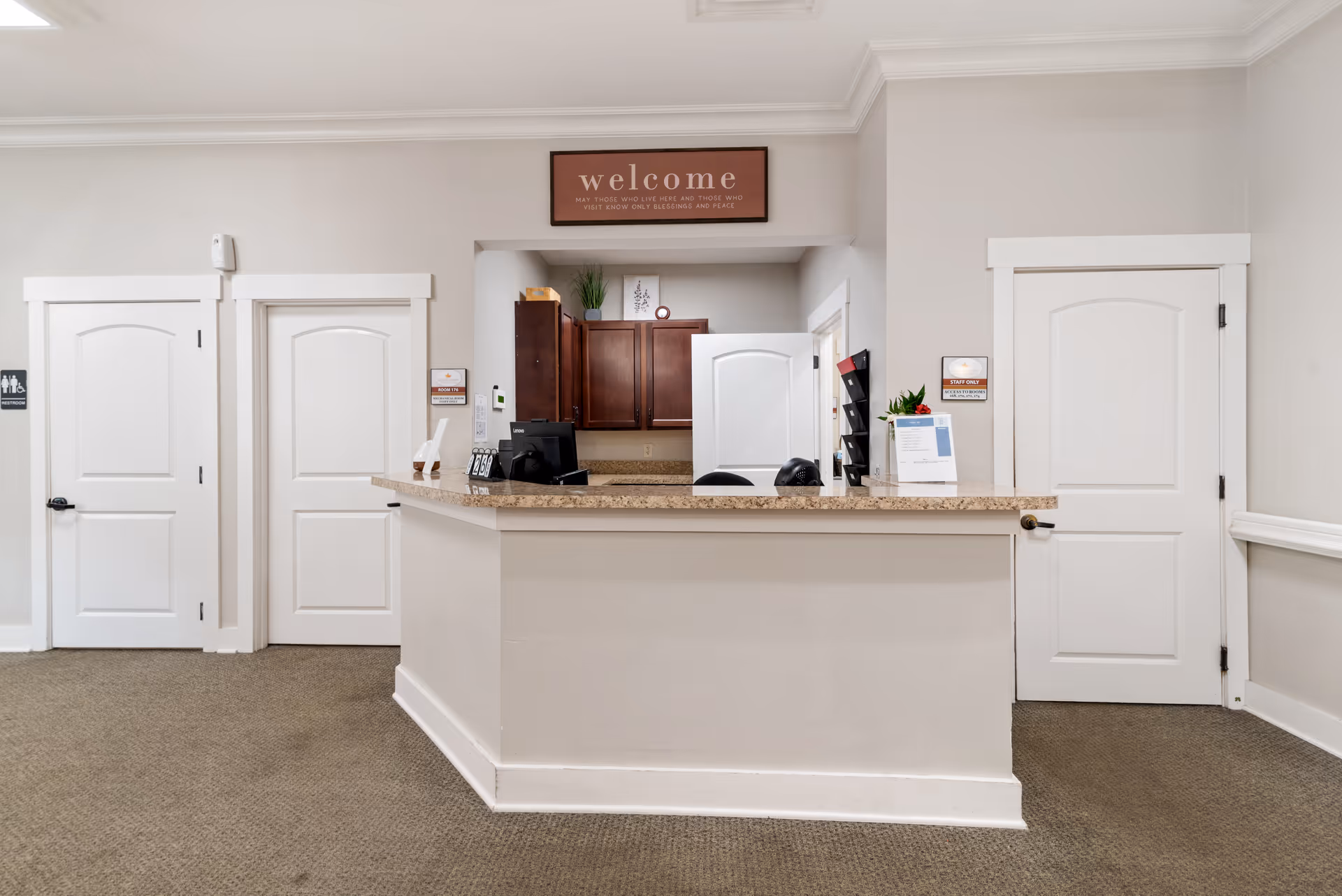 Reception desk in a neutral-colored lobby with a 'welcome' sign above, three white doors, and office equipment on the counter.