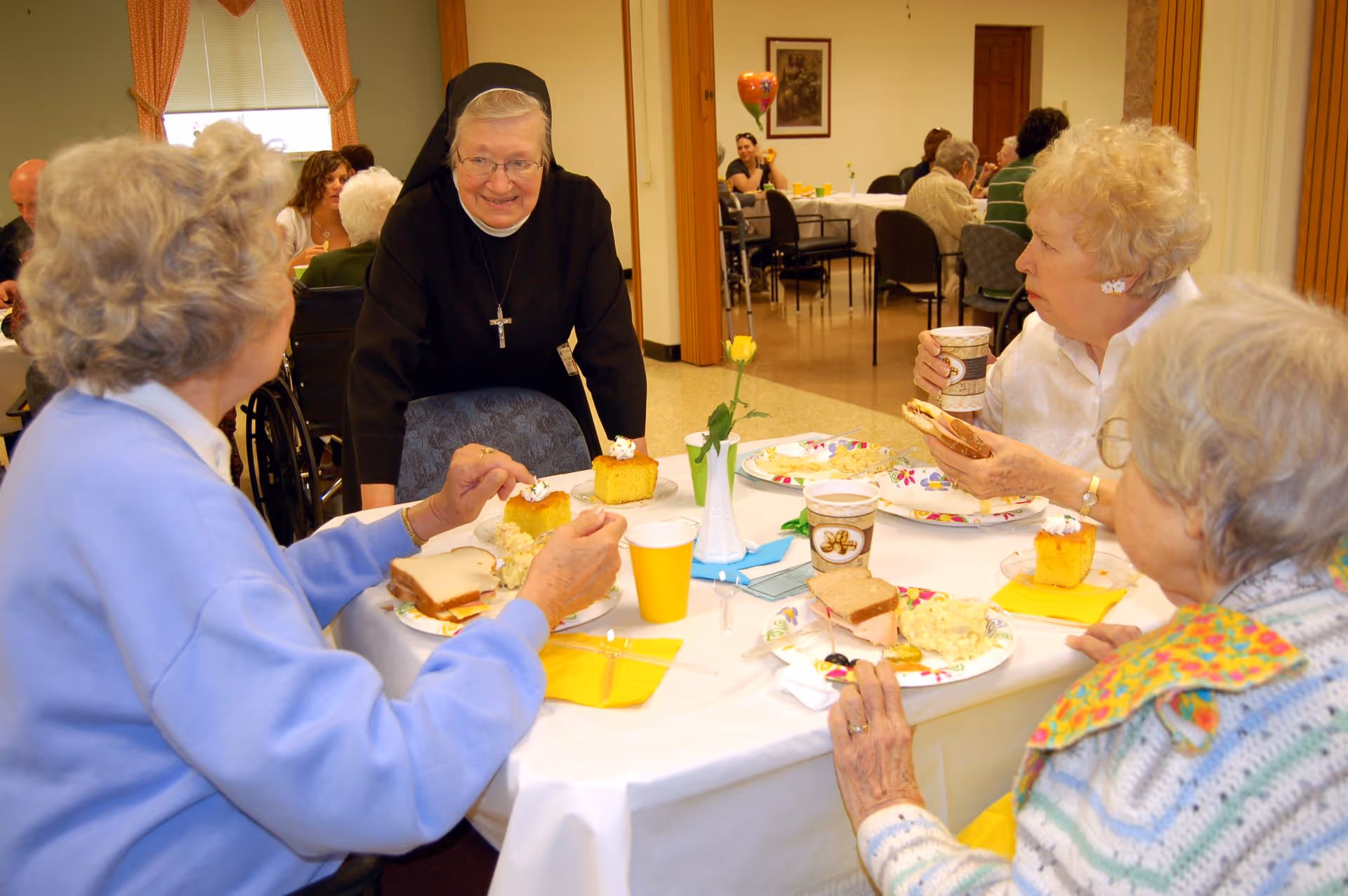A group of elderly women sitting around a table enjoying a meal together in a dining room. A nun is standing and smiling at the women. The table is set with plates of food, cups, and a small vase with a yellow flower. Other people are visible in the background also seated at tables.