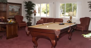 A cozy common room with a pool table in the center, surrounded by comfortable armchairs and a sofa. The room has large windows letting in natural light, a wooden cabinet with a painting above it, and decorative plants and lamps.