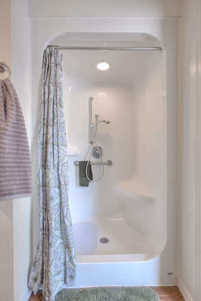 A white shower stall with a patterned curtain, handheld showerhead, grab bar, and towels.