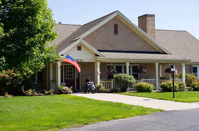 A single-story brick building with a covered porch, an American flag mounted near the entrance, and a person sitting in a wheelchair on the porch. The building is surrounded by green grass, bushes, and hanging flower baskets under a clear sky.