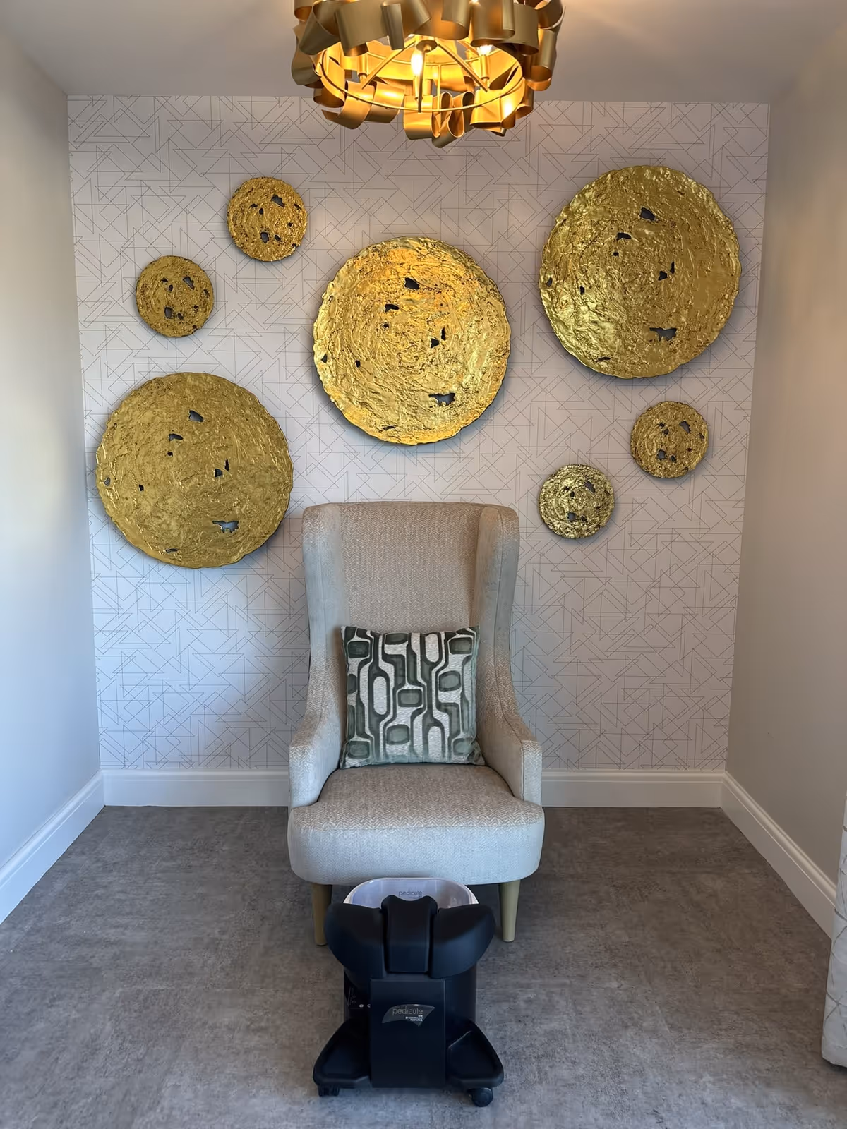 Upholstered armchair with a patterned pillow beneath gold circular wall sculptures and a foot massager in a small decorated alcove.