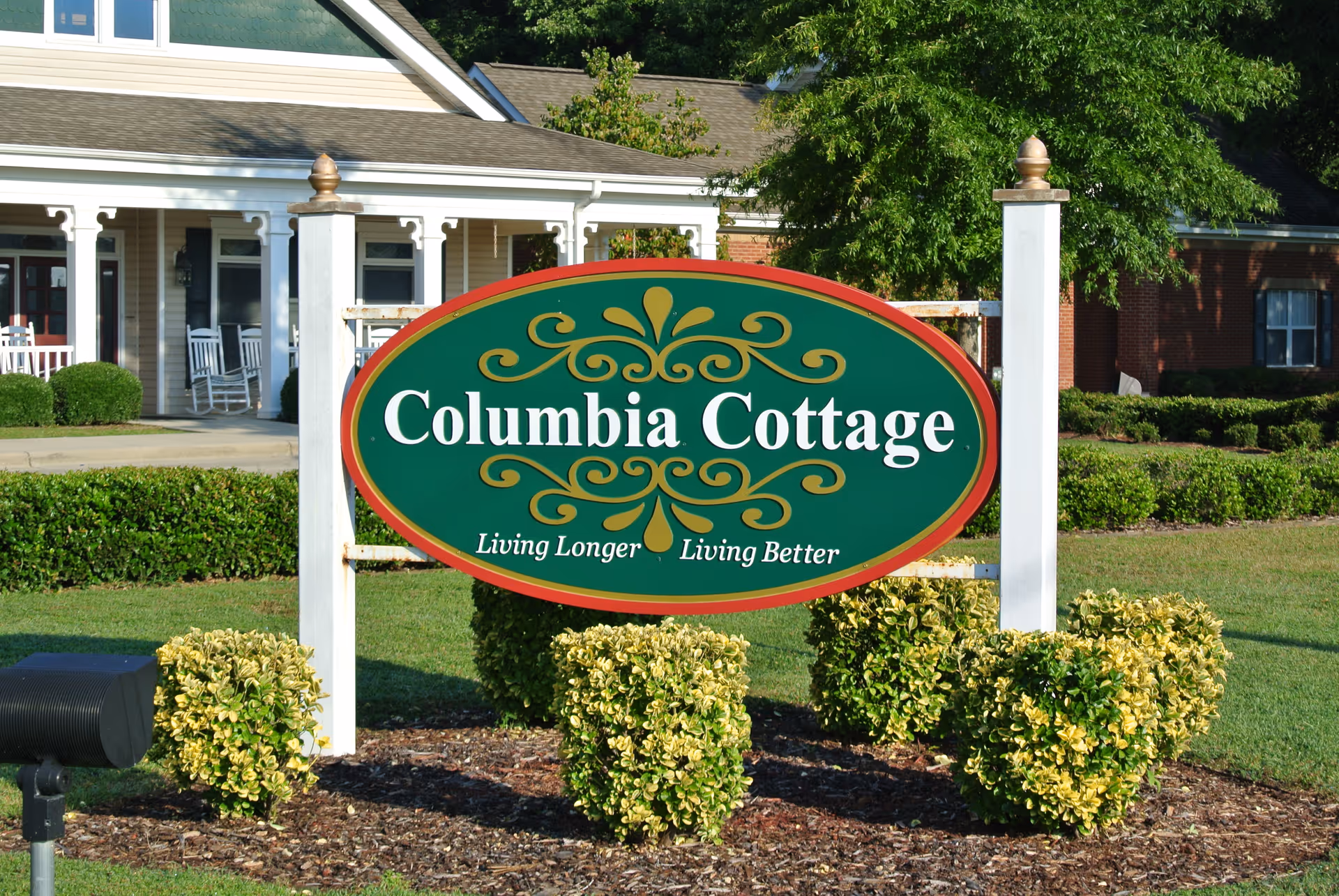 Outdoor view of a green and red sign for Columbia Cottage with the slogan 'Living Longer Living Better' in front of a building with white pillars, rocking chairs on the porch, and well-maintained bushes and lawn.