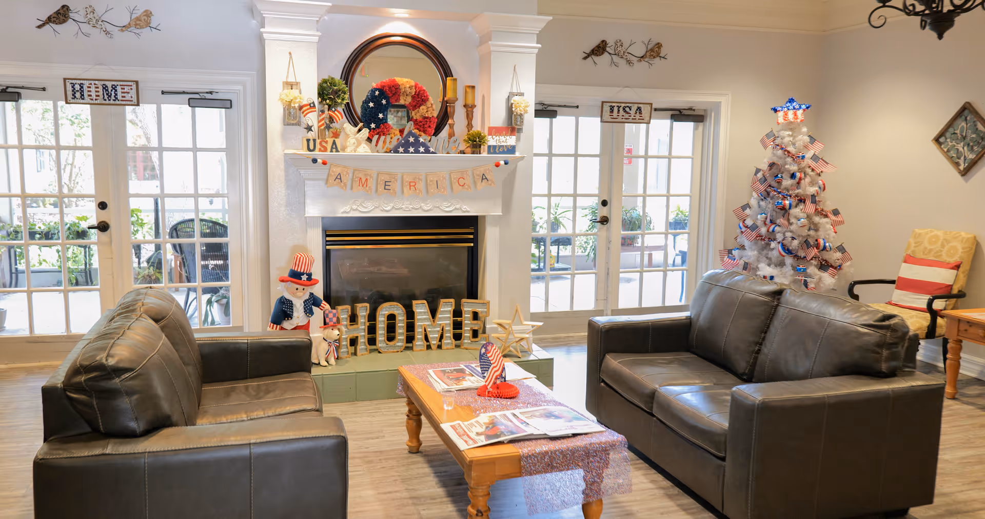A cozy living room with two dark leather sofas facing each other and a wooden coffee table in between. The room features a fireplace decorated with patriotic American-themed items including a wreath, candles, and a banner spelling AMERICA. There is a white Christmas tree adorned with American flags and red, white, and blue decorations. French doors with glass panes lead outside, and there are additional patriotic decorations on the walls and furniture.