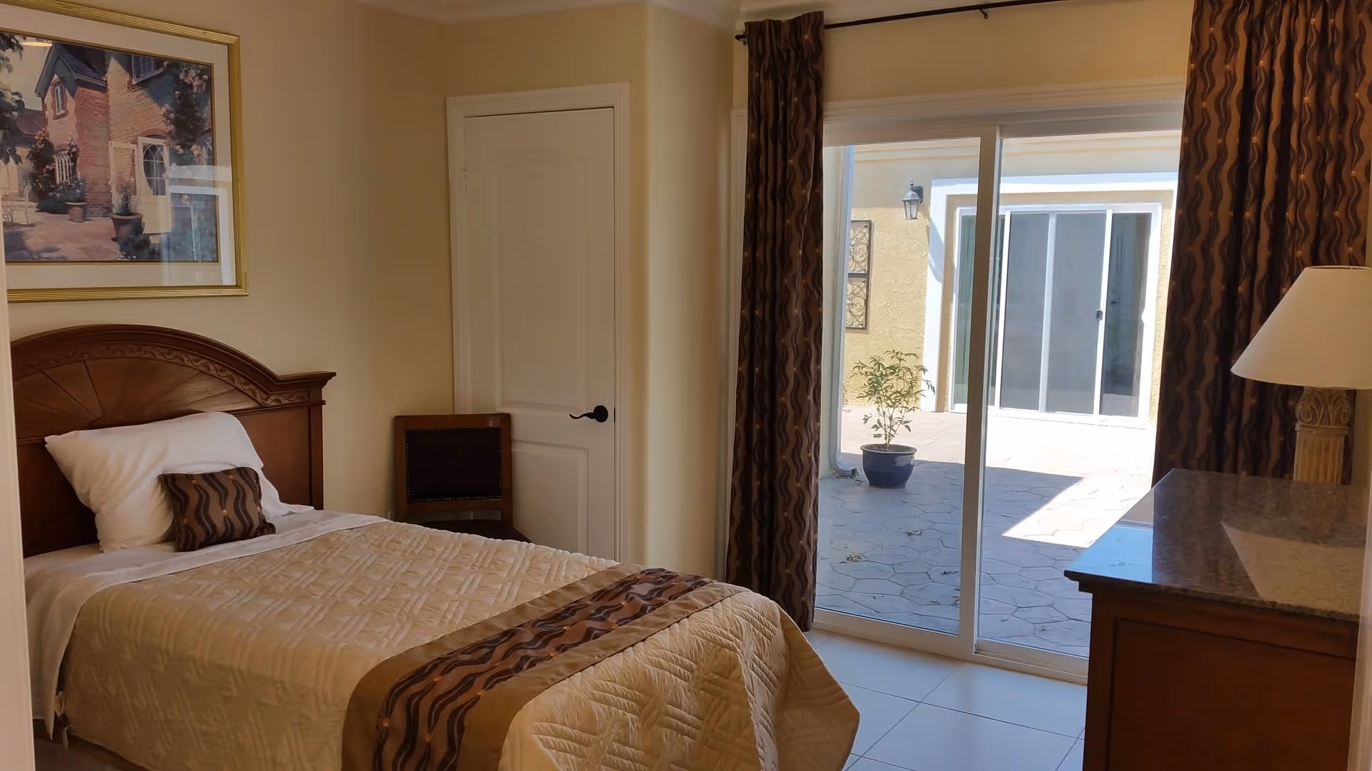 A bedroom with a single bed featuring a beige quilt and a decorative pillow. There is a wooden headboard, a chair next to a closed white door, and a dresser with a lamp on it. Sliding glass doors with patterned curtains open to an outdoor patio area with a potted plant.