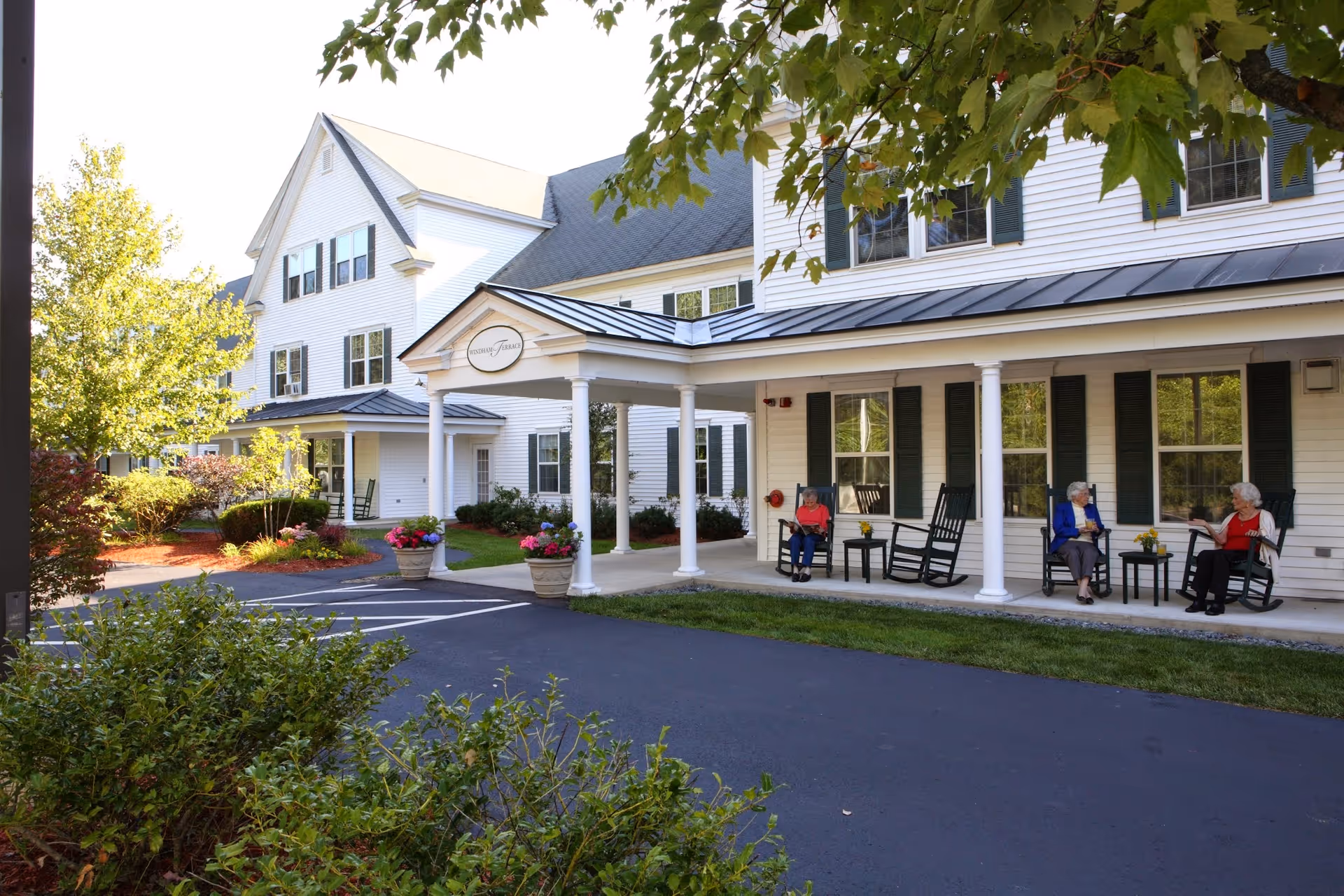 Exterior view of Windham Terrace, a white multi-story senior living facility with black shutters and a covered entrance. Three elderly women are sitting on rocking chairs on the porch, engaged in conversation. The area is surrounded by greenery and potted flowers, with a paved driveway in front.