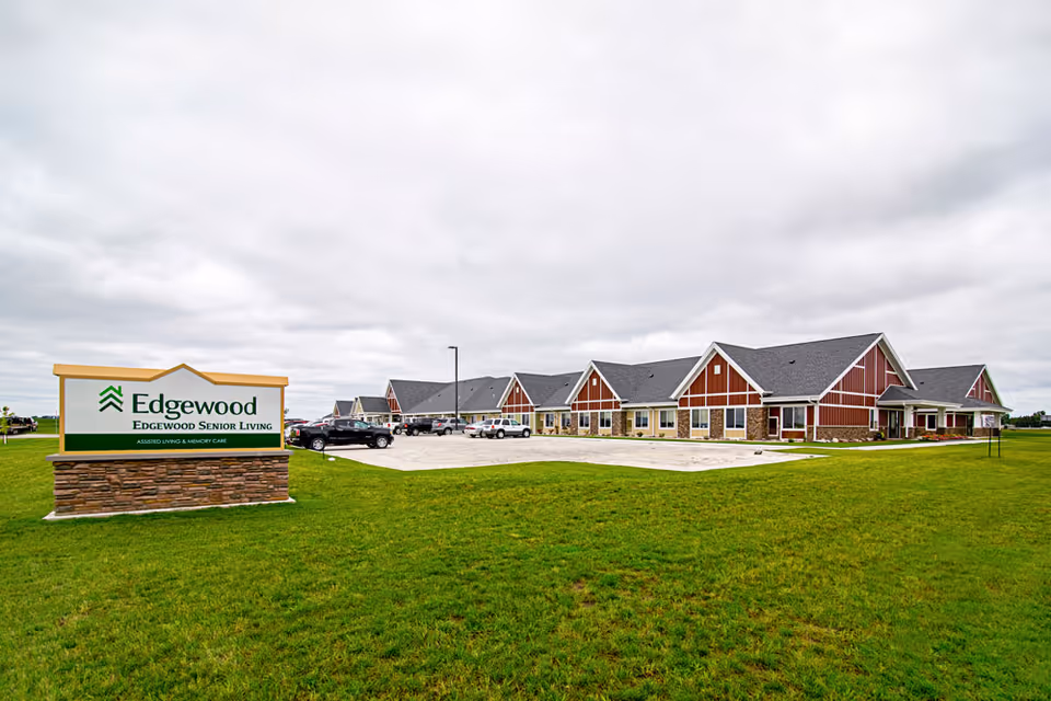 Exterior view of Edgewood Senior Living facility with a large green lawn in the foreground, a parking lot with several cars, and a long building with red and beige siding and multiple peaked roofs under a cloudy sky.