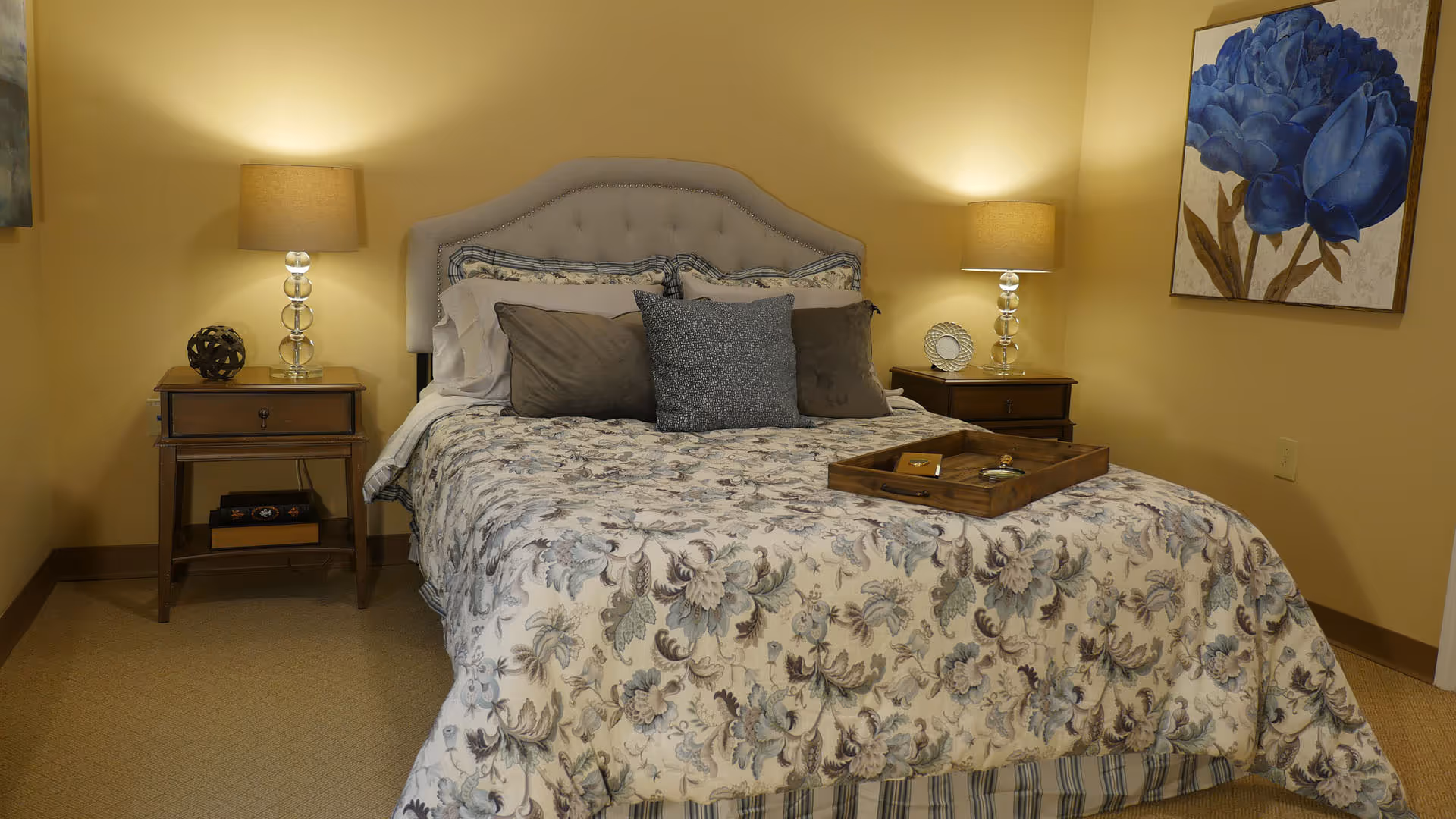 A neatly made bed with a floral patterned bedspread flanked by two nightstands and lamps in a warmly lit bedroom.