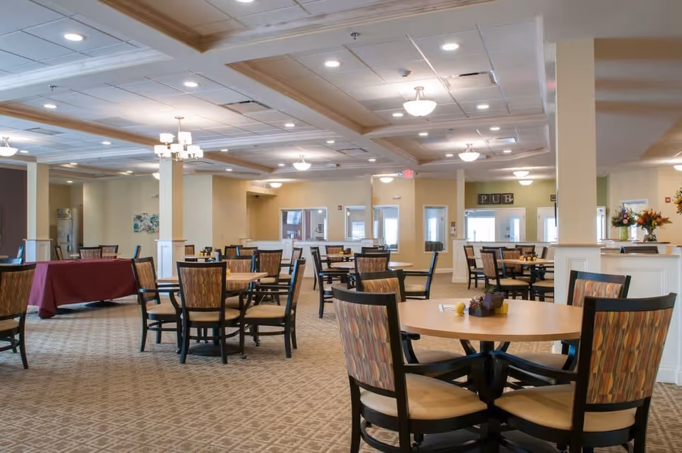 Spacious dining room with multiple round tables and chairs arranged neatly. The room has a patterned carpet, beige walls, and a ceiling with recessed lighting and chandeliers. There is a table covered with a maroon cloth on the left side and a sign that reads 'PUB' on the far wall.