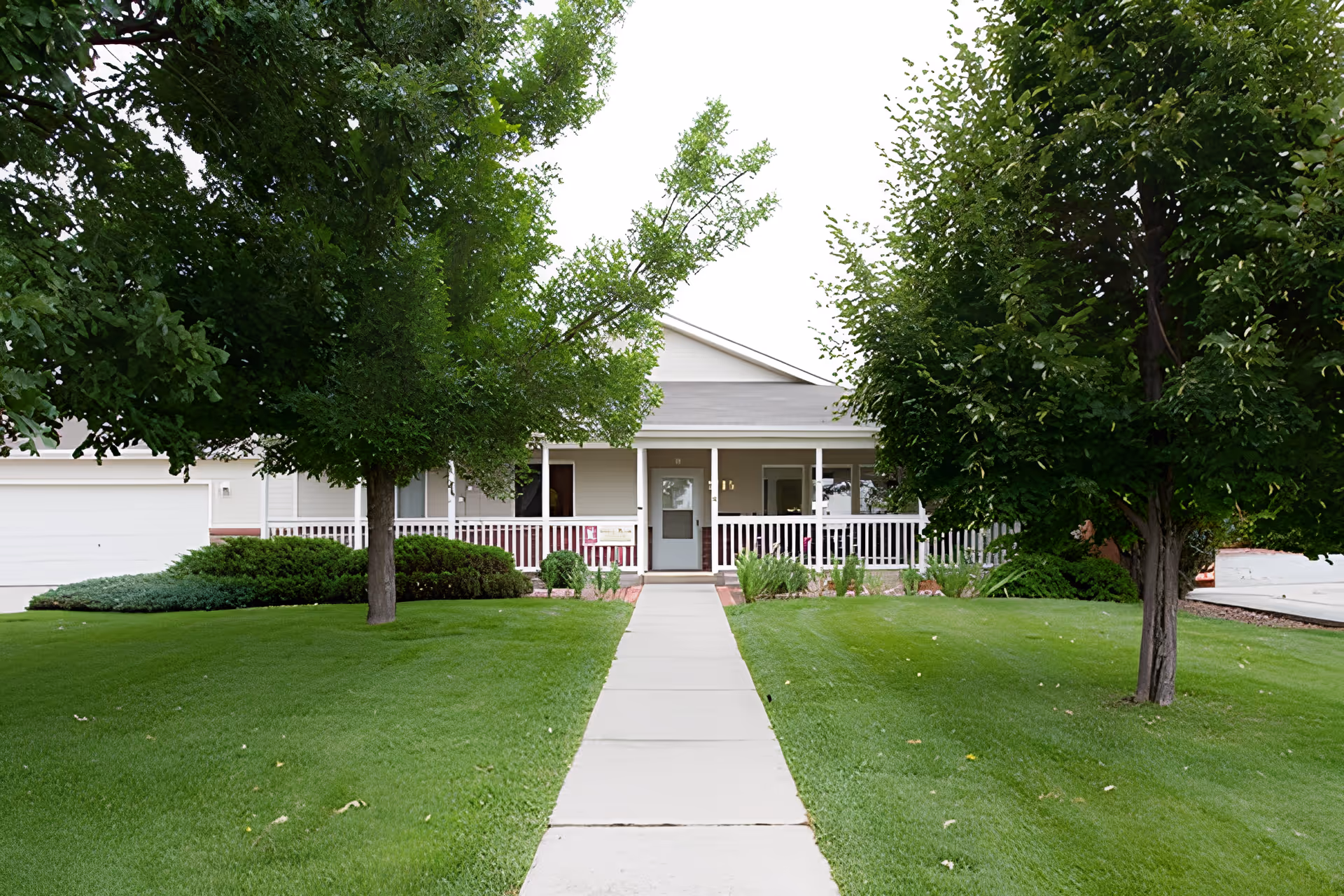 Front exterior view of a single-story building with a covered porch, white railing, and a concrete walkway leading to the entrance. The building is surrounded by well-maintained green grass, bushes, and two large trees on either side of the walkway.