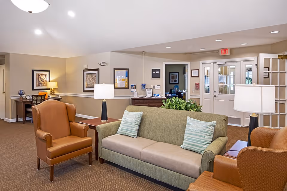 A well-lit senior living facility lounge with sofas, armchairs, side tables, and a reception area in the background.