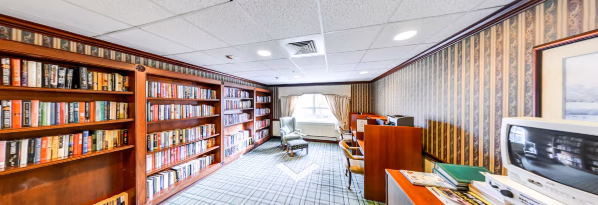 A cozy library room with large wooden bookshelves filled with books along the left wall, a window with curtains at the far end, two armchairs near the window, and a row of wooden desks with chairs and desktop computers on the right side. The room has patterned wallpaper and a carpeted floor with a checkered design.