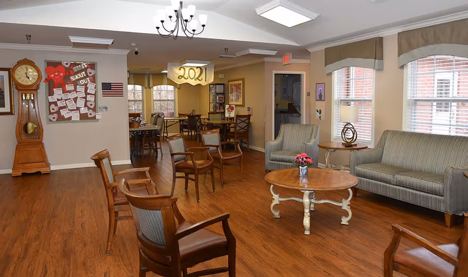 A cozy assisted living common area with wooden flooring, several wooden chairs arranged around a round wooden coffee table with a small flower vase on it. There is a striped sofa and an armchair near two windows with blinds and valances. A grandfather clock stands against the wall near a bulletin board decorated with heart-shaped notes and an American flag. In the background, there are more tables and chairs in a dining area, with a hanging decoration displaying '2021'.