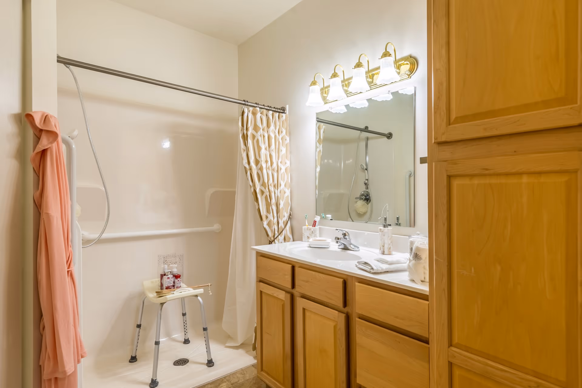 A bathroom with a walk-in shower featuring a shower chair and a grab bar. There is a shower curtain with a geometric pattern partially drawn. A pink robe hangs on a hook near the shower. The vanity has a large mirror with five light fixtures above it, a sink, and wooden cabinets below. Various toiletries and towels are placed on the countertop.