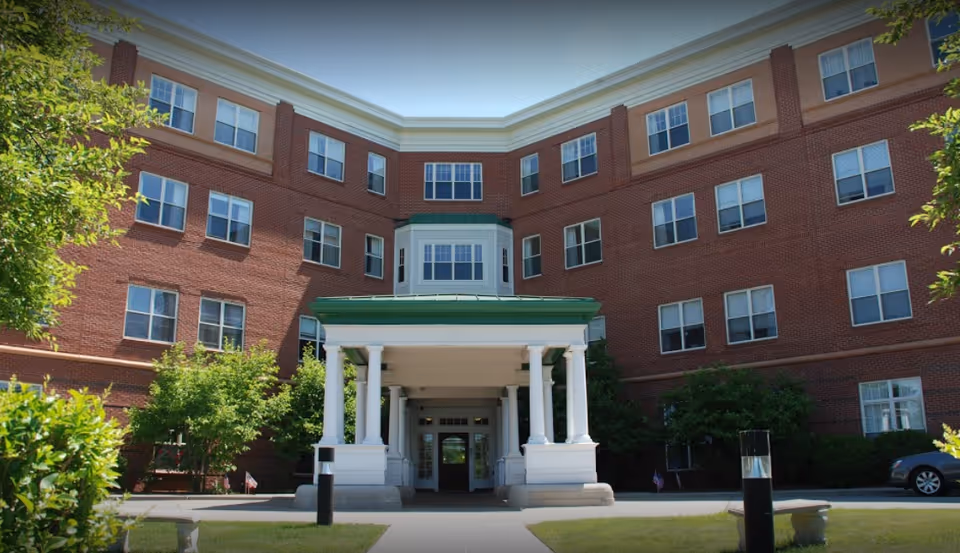 Front entrance of a four-story red brick residential building featuring a white-columned portico with a green roof and landscaped grounds.