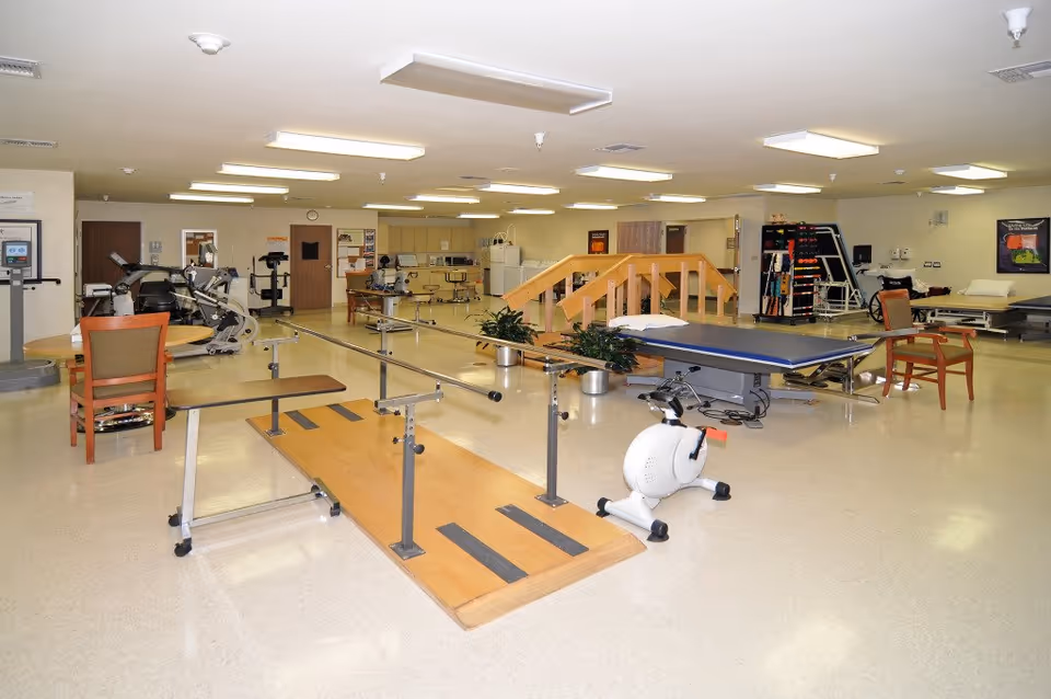 A spacious physical therapy room with exercise equipment including parallel bars for walking practice, stationary bikes, a treatment table, wooden stairs for rehabilitation, chairs, and various therapy tools. The room is well-lit with ceiling lights and has a clean, polished floor.