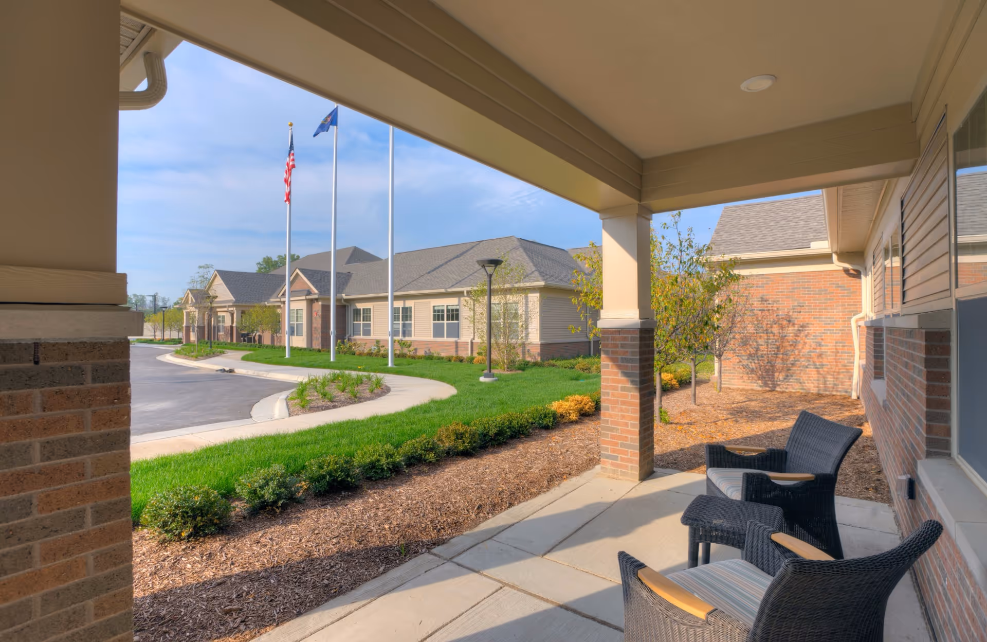 Covered porch with wicker chairs overlooking the landscaped entrance and flagpoles of a senior living facility.