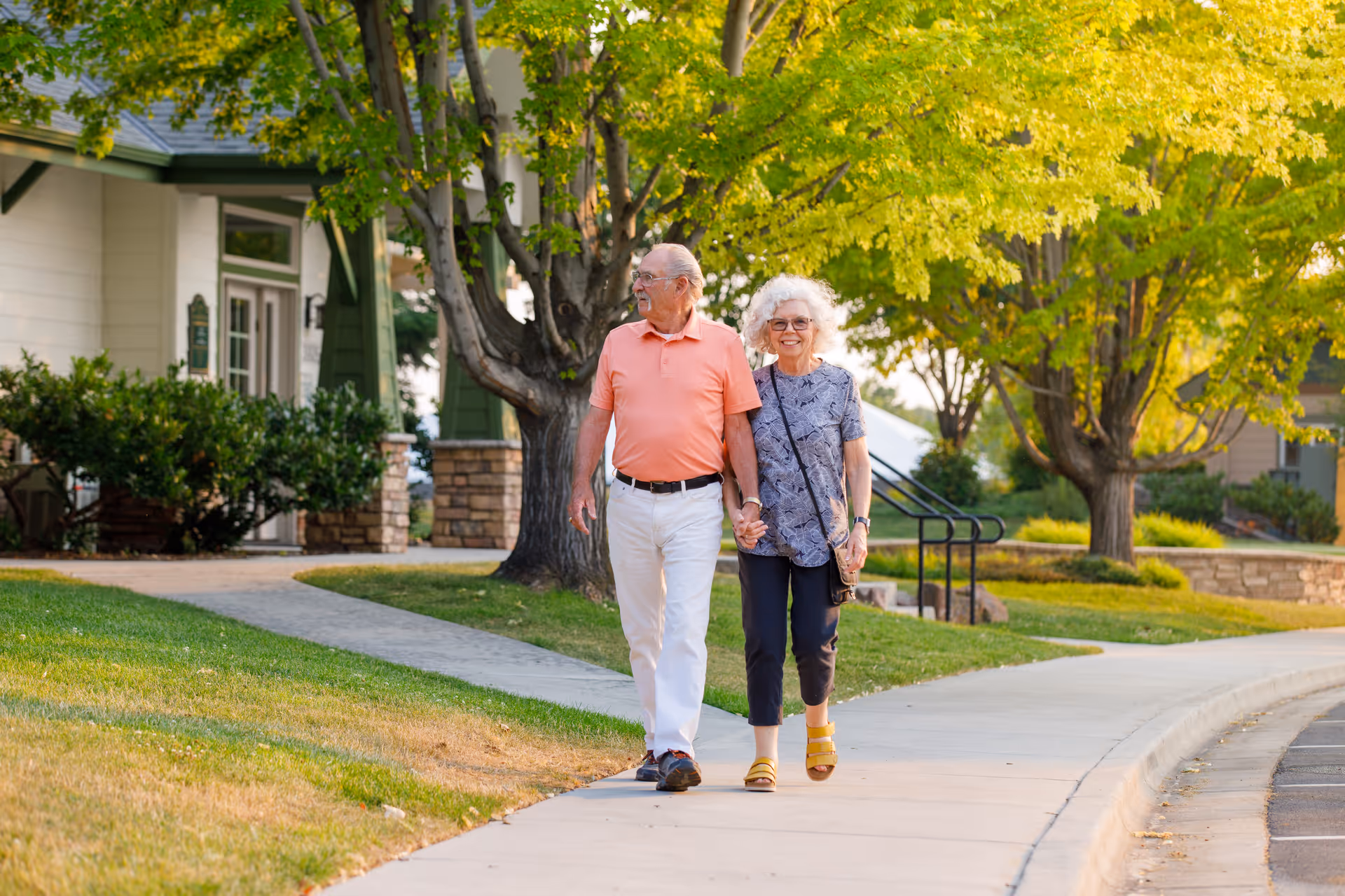 An elderly couple holding hands and walking on a sidewalk in a residential area with green trees and grass around them. The man is wearing a coral polo shirt and white pants, and the woman is wearing a patterned blouse and dark pants with yellow sandals. They appear happy and relaxed.