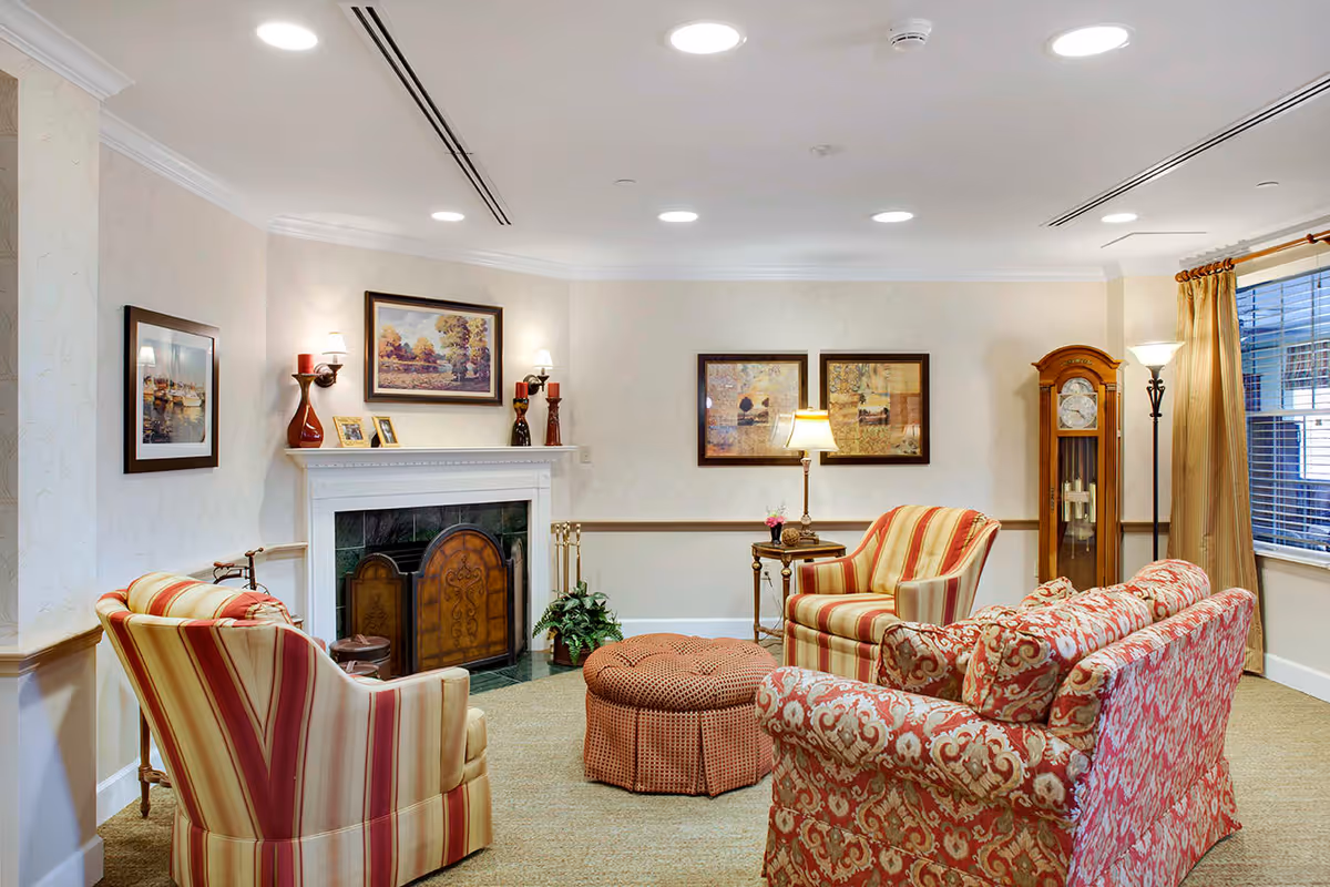 A cozy living room with patterned upholstered armchairs and a sofa arranged around a round ottoman. The room features a white fireplace with decorative items on the mantel, framed artwork on the walls, a grandfather clock, and a window with beige curtains. The lighting includes ceiling recessed lights and floor lamps.