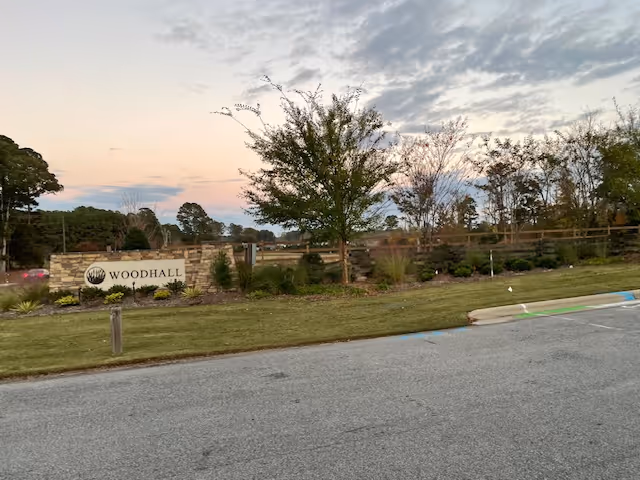 Entrance sign reading "Woodhall" with landscaped lawn, trees, and a roadway in the foreground at dusk.