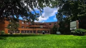 Exterior view of a three-story brick senior living facility named St Cloud Carefree Living, with large windows and a green lawn in the foreground under a partly cloudy blue sky.
