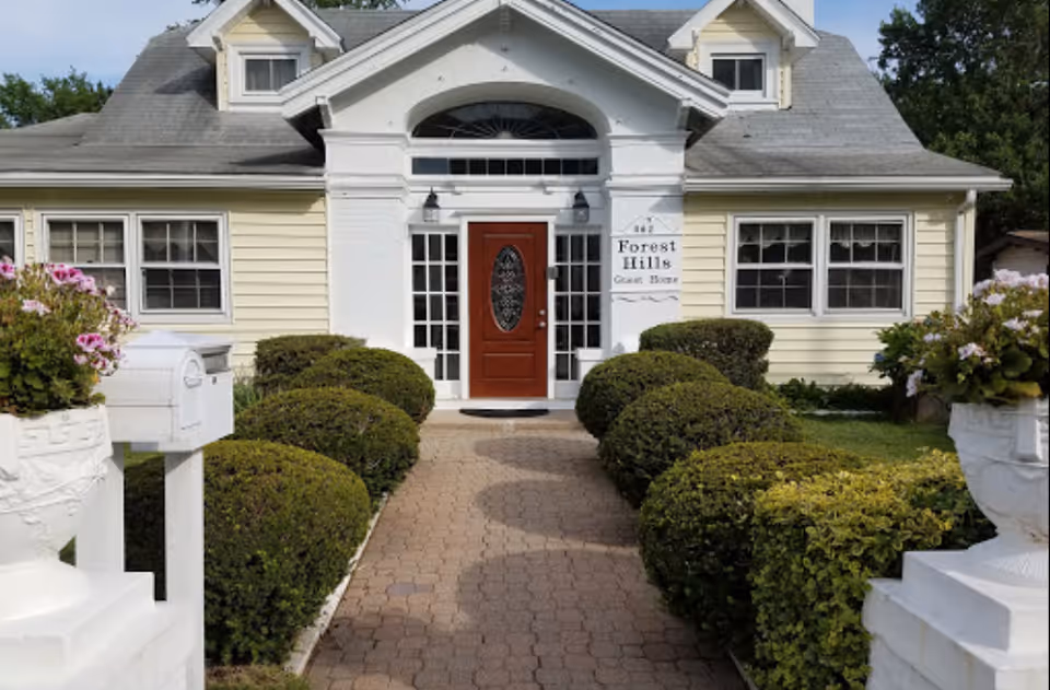 Front entrance of a light-yellow guest home with a red door, manicured shrubs, and a 'Forest Hills Guest Home' sign.