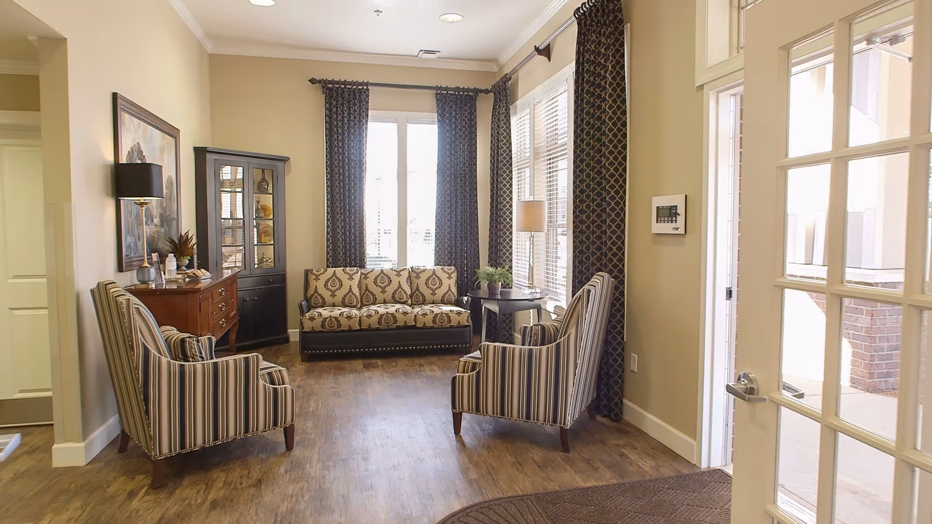 A cozy sitting area in a senior living facility with two striped armchairs facing a patterned sofa. The room has large windows with dark patterned curtains, a wooden floor, a small side table with a lamp and plant, a wooden cabinet with decorative items, and a glass-paneled door leading outside.