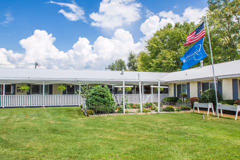 Exterior view of a single-story senior living facility building with a white roof and white railing. There is a well-maintained green lawn and garden area with shrubs and flowers in front of the building. Two flags, one American and one blue flag, are flying on a flagpole near the building. Trees and a partly cloudy sky are visible in the background.
