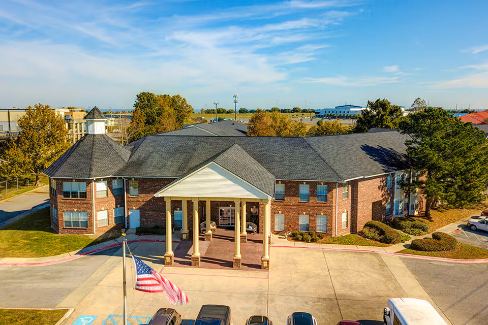 Front exterior of a two-story brick senior living building with a columned entrance, parking lot, and American flag.