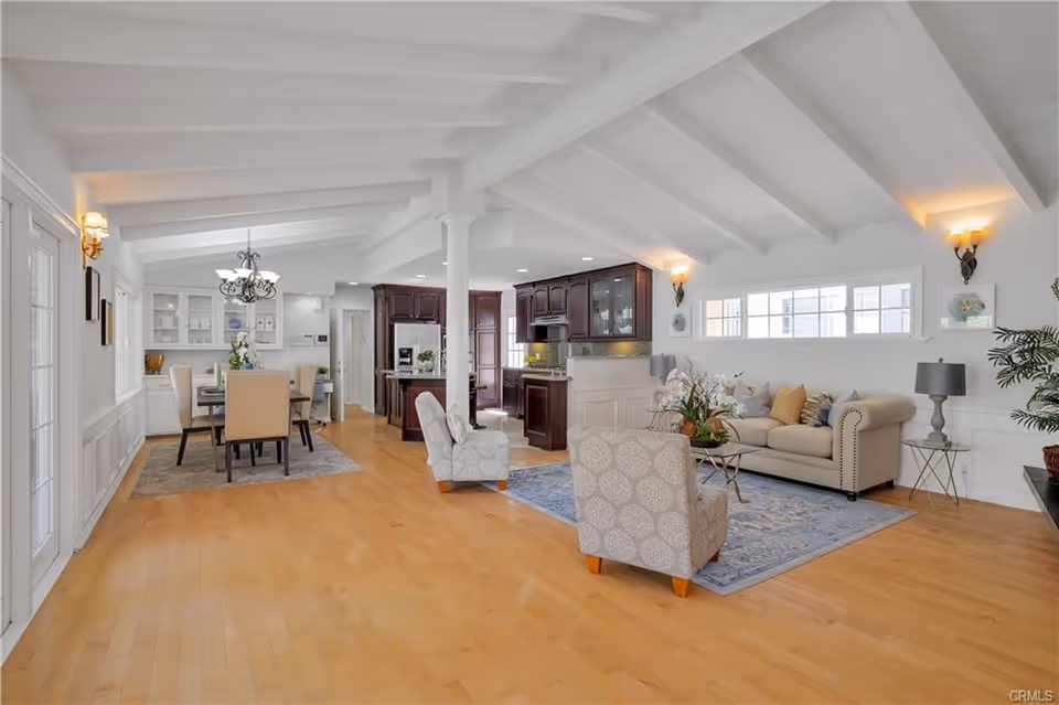 Bright and spacious open-concept living and dining area with light wood flooring, white vaulted ceiling with exposed beams, beige sofa with decorative pillows, two patterned armchairs, a glass coffee table with a floral arrangement, a dining table with six beige upholstered chairs under a chandelier, and a kitchen with dark wood cabinets in the background.
