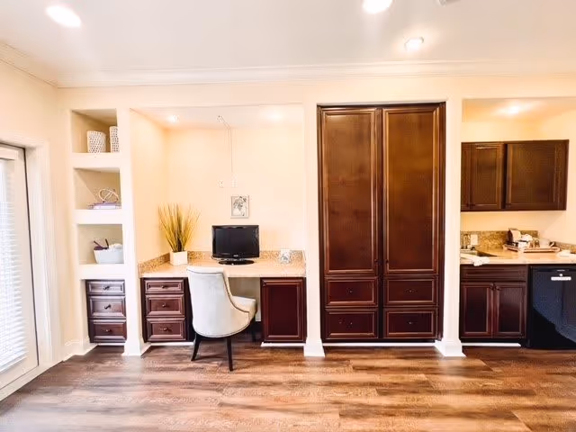 Interior view of a room featuring a built-in desk area with a computer monitor and a cushioned chair. To the right, there are dark wood cabinets and a kitchen countertop with a sink and dishwasher. The floor is wooden, and the walls are light-colored with recessed lighting in the ceiling.