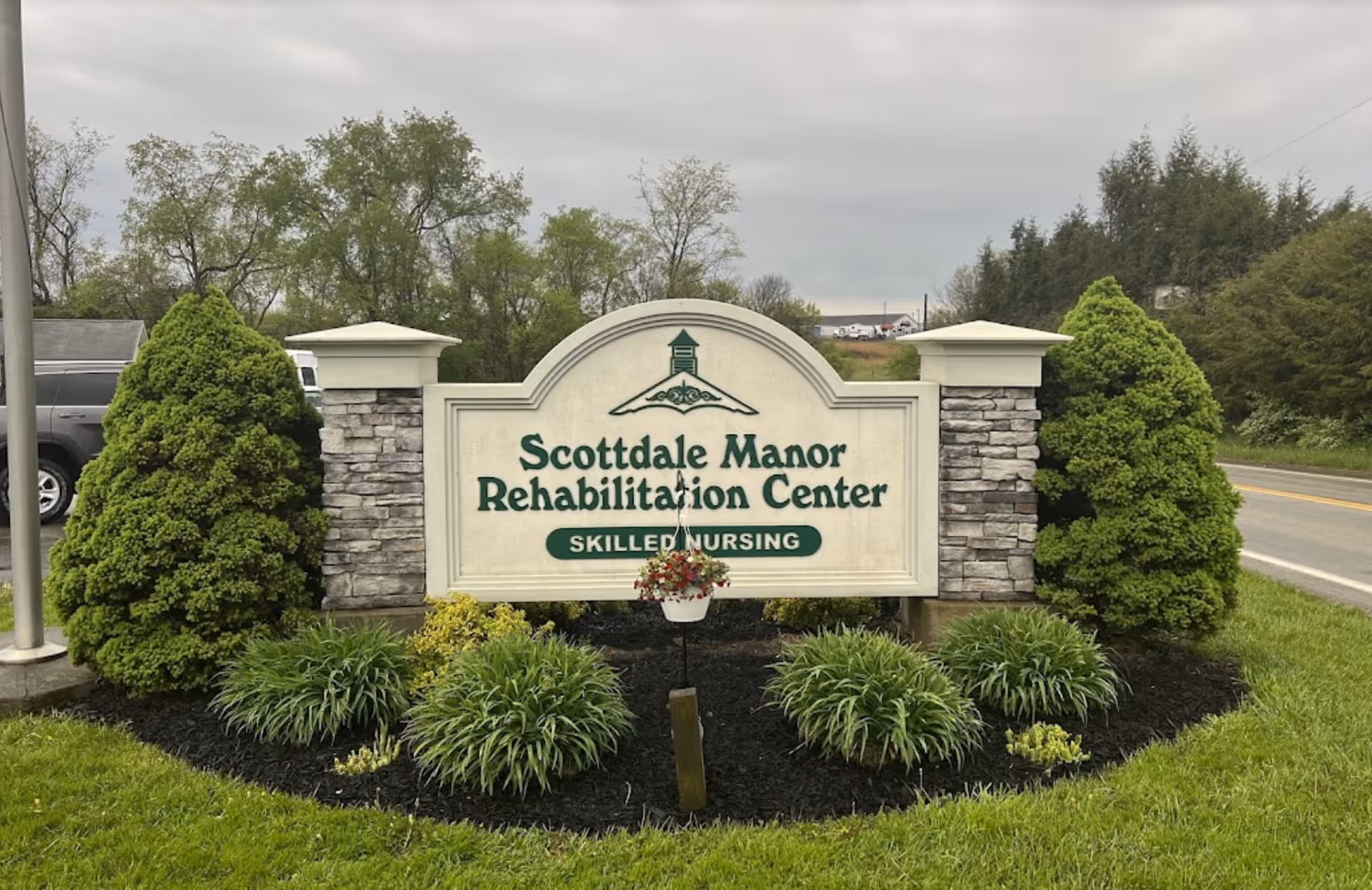 Outdoor view of a sign for Scottdale Manor Rehabilitation Center Skilled Nursing, surrounded by neatly trimmed bushes and plants, with a road and trees in the background under a cloudy sky.
