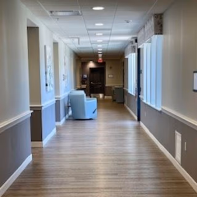A long, well-lit hallway in a senior living facility with light wood flooring, beige and gray walls, and a light blue armchair positioned along the left side. The hallway has several windows on the right side and framed artwork on the left wall, leading to a dark door at the end.