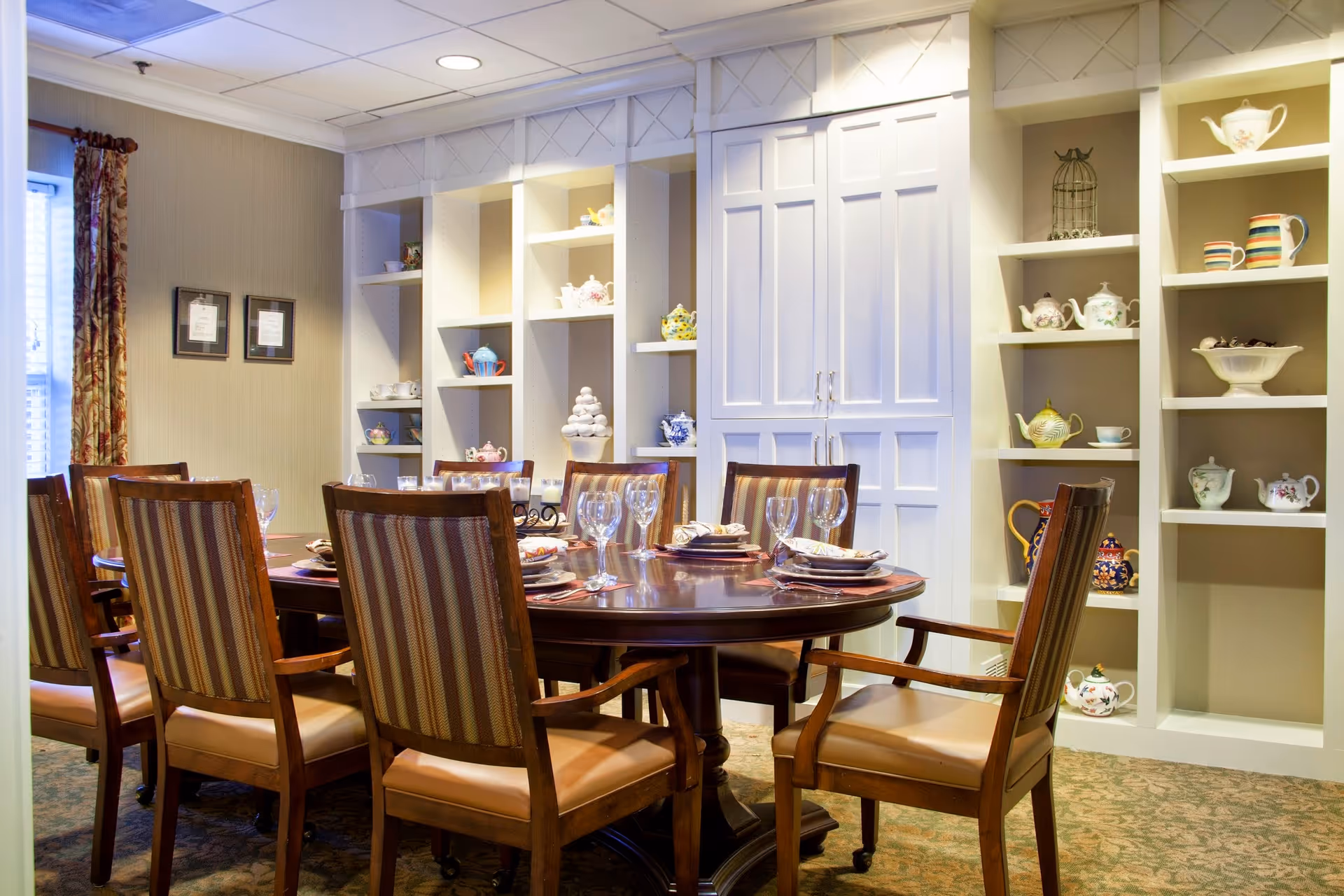 A formal dining room with a round wooden table set for six people with plates, napkins, and wine glasses. The chairs have wooden frames with striped upholstery. The room features built-in white shelves displaying various teapots and decorative items, and there are two framed pictures on the wall near a window with patterned curtains.