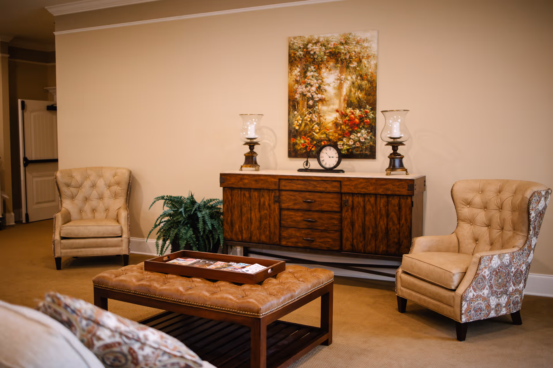 A cozy living room area with two beige tufted armchairs, a wooden sideboard with three drawers and two cabinets, a decorative clock, two candle holders, a green potted plant, and a colorful floral painting hanging on the wall. In the foreground, there is a cushioned ottoman with a wooden tray holding magazines.