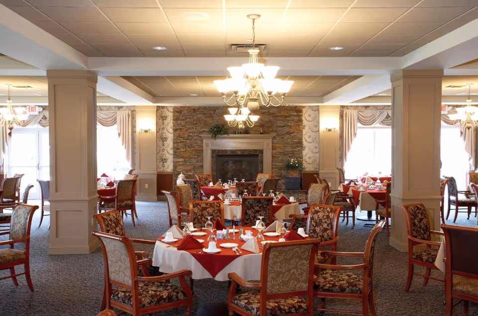 A spacious dining room with multiple round tables covered with white and red tablecloths, set with napkins, cups, and silverware. The room features floral upholstered chairs, a stone fireplace in the background, chandeliers hanging from the ceiling, and large windows with draped curtains allowing natural light to enter.