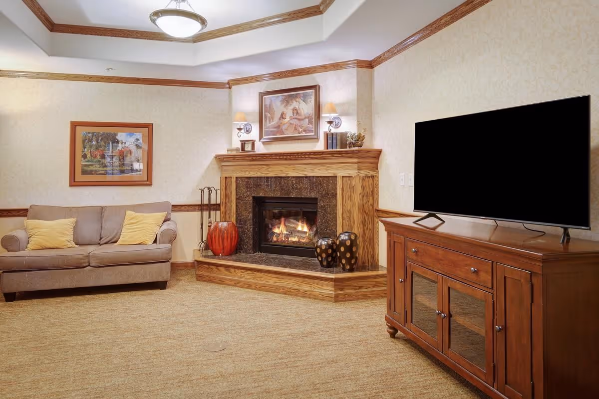 A cozy living room with a beige couch adorned with two yellow pillows, a wooden fireplace with a fire burning inside, decorative vases on the hearth, framed artwork above the fireplace, and a large flat-screen TV on a wooden cabinet.