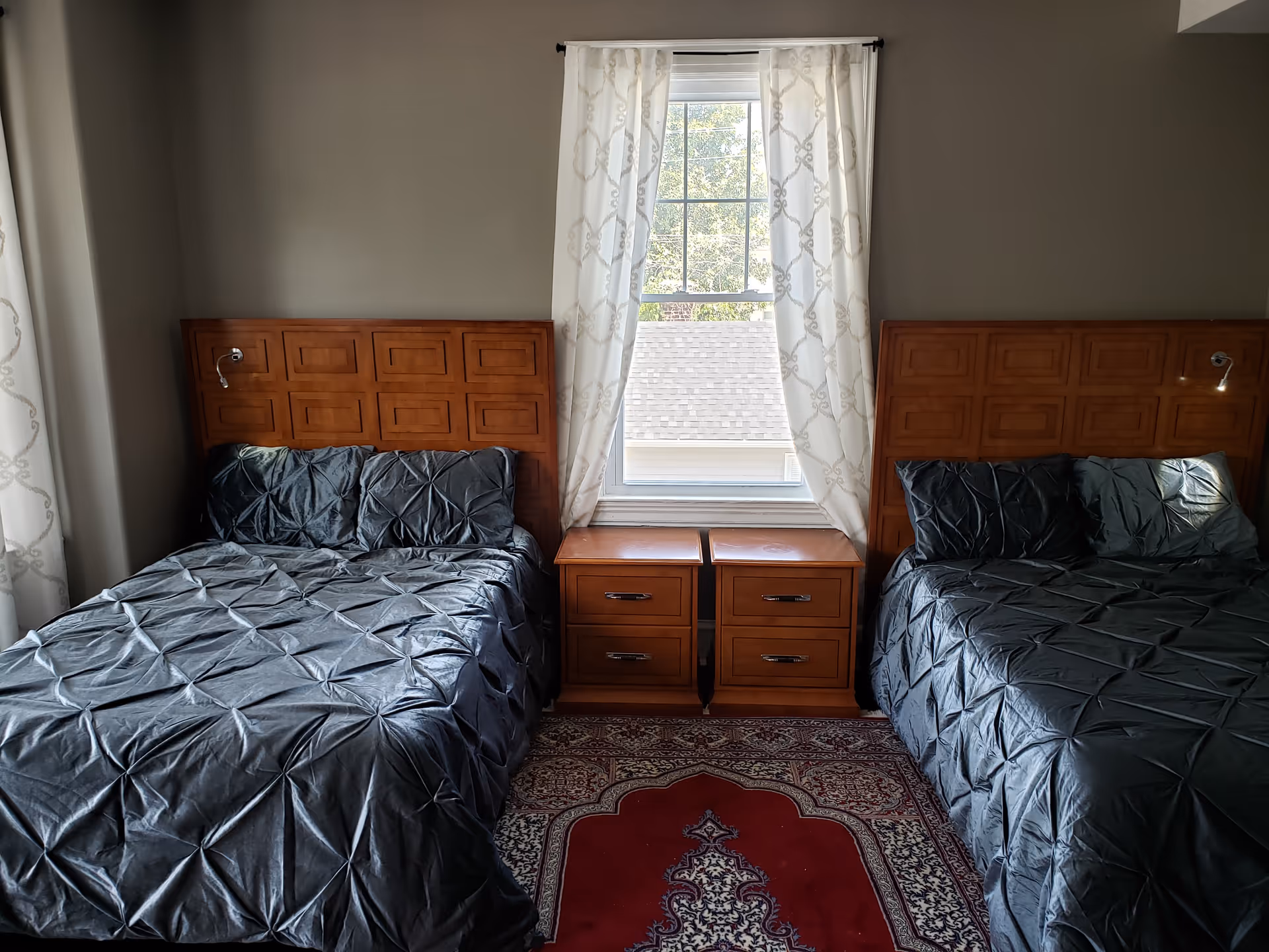 Two beds with dark bedding and wooden headboards flanking a window with curtains and matching nightstands between them.
