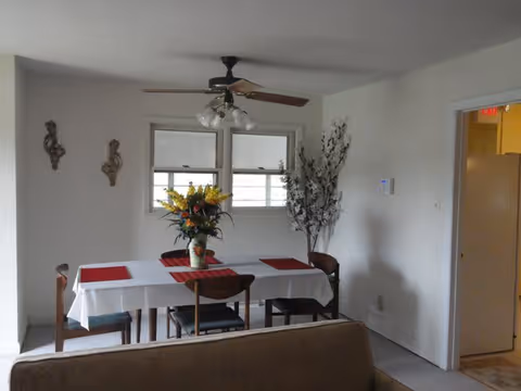 A dining area with a rectangular table covered with a white tablecloth and four wooden chairs. The table has a vase with a colorful flower arrangement and red placemats. There is a ceiling fan with lights above the table, two windows on the wall behind, and a tall decorative plant in the corner. Part of a couch is visible in the foreground and a doorway leading to another room is on the right.