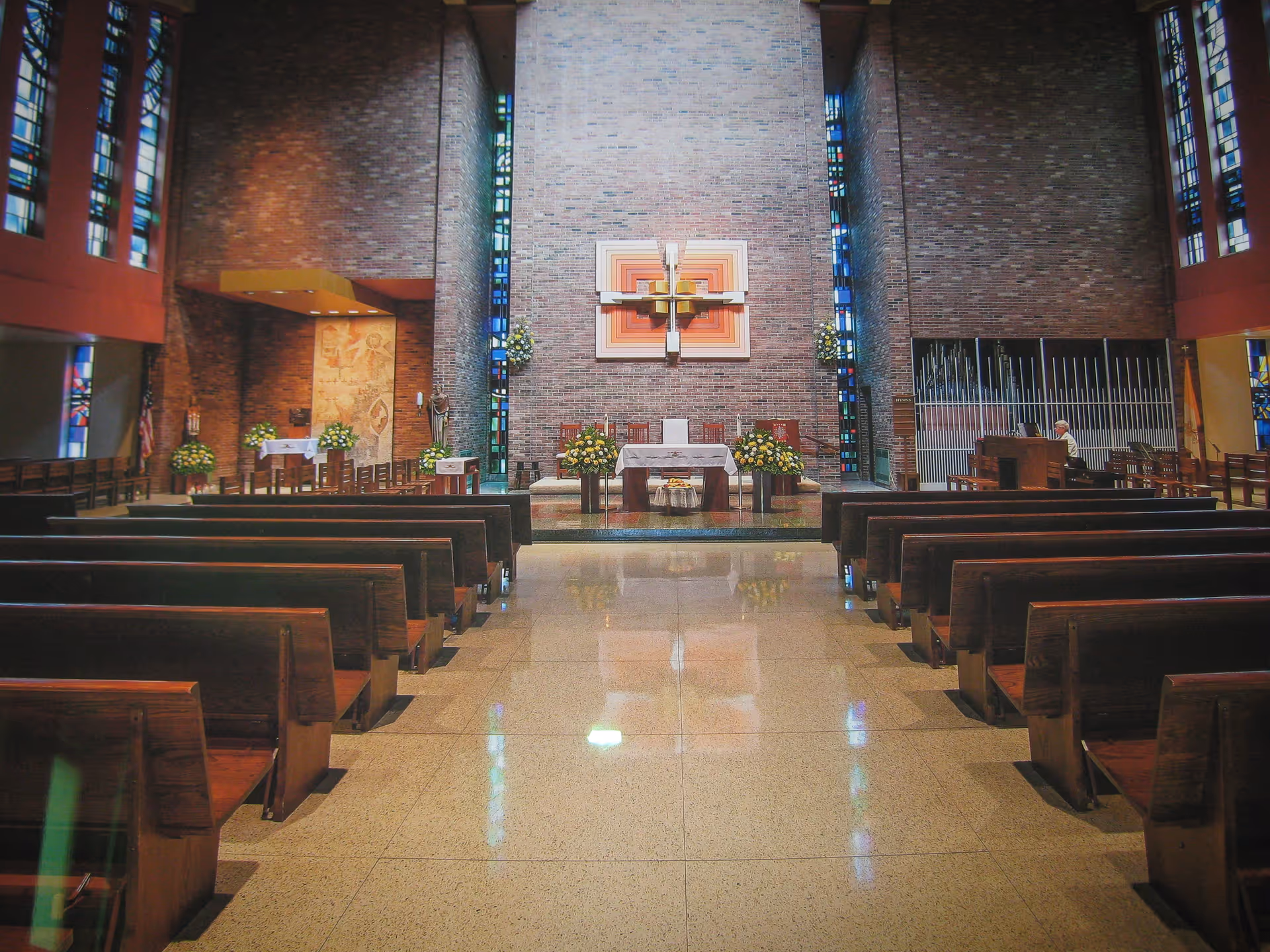Interior view of a chapel with wooden pews arranged in rows facing an altar. The altar is decorated with flower arrangements and a large cross mounted on a brick wall behind it. Stained glass windows flank the altar area, and there is a person seated at an organ on the right side. The floor is polished and reflective.