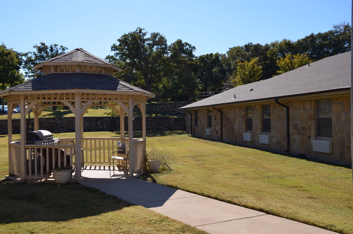 Outdoor area of Green Oaks Nursing and Rehabilitation featuring a wooden gazebo with a grill and a chair inside, surrounded by a grassy lawn and a stone building with multiple windows in the background under a clear blue sky.