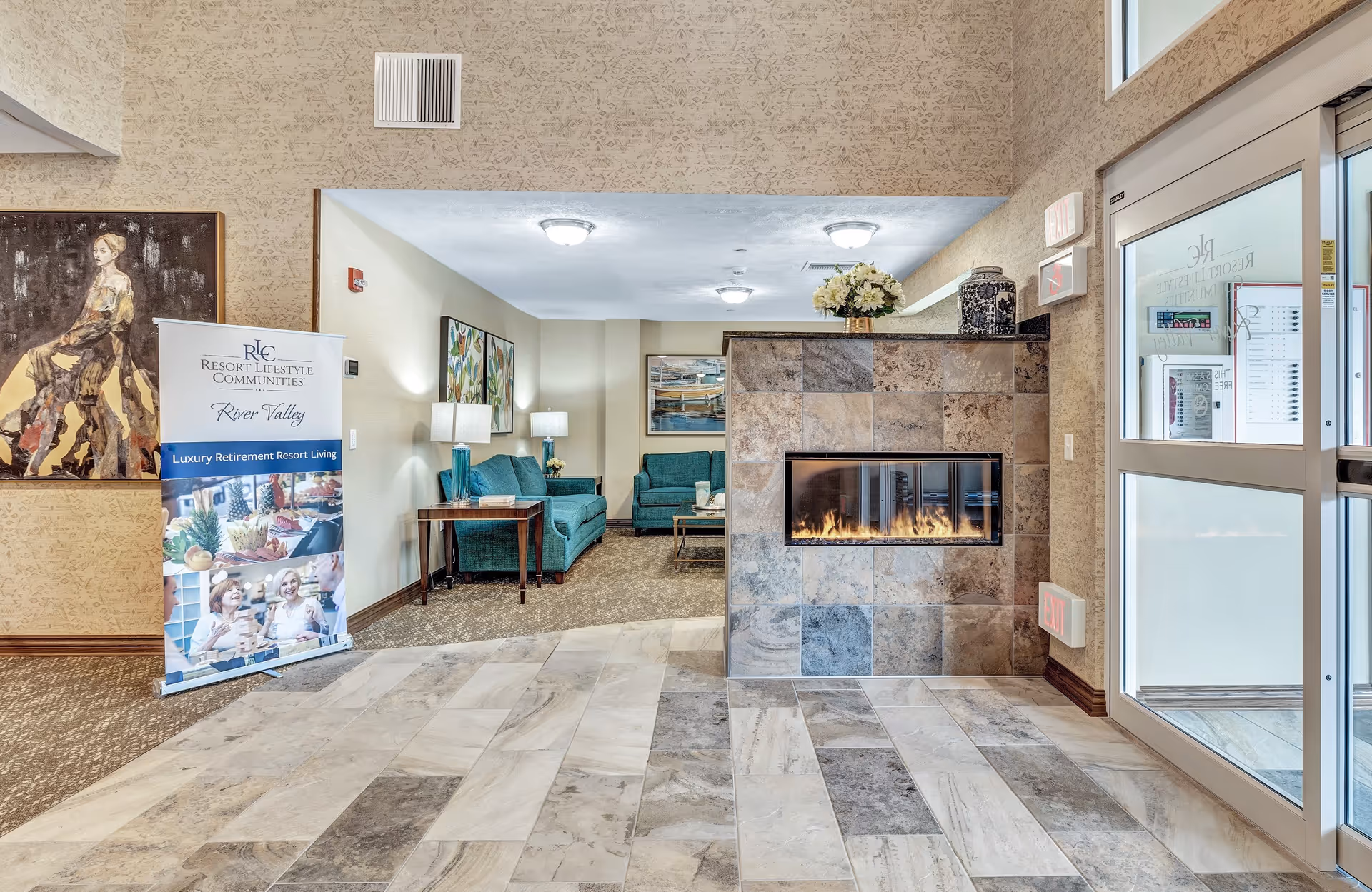 Interior view of a retirement community lobby area featuring a modern tiled fireplace with a fire burning, teal sofas and side tables with lamps in the background, a large painting on the wall, and a standing banner that reads Resort Lifestyle Communities River Valley Luxury Retirement Resort Living. There is a glass entrance door on the right side.