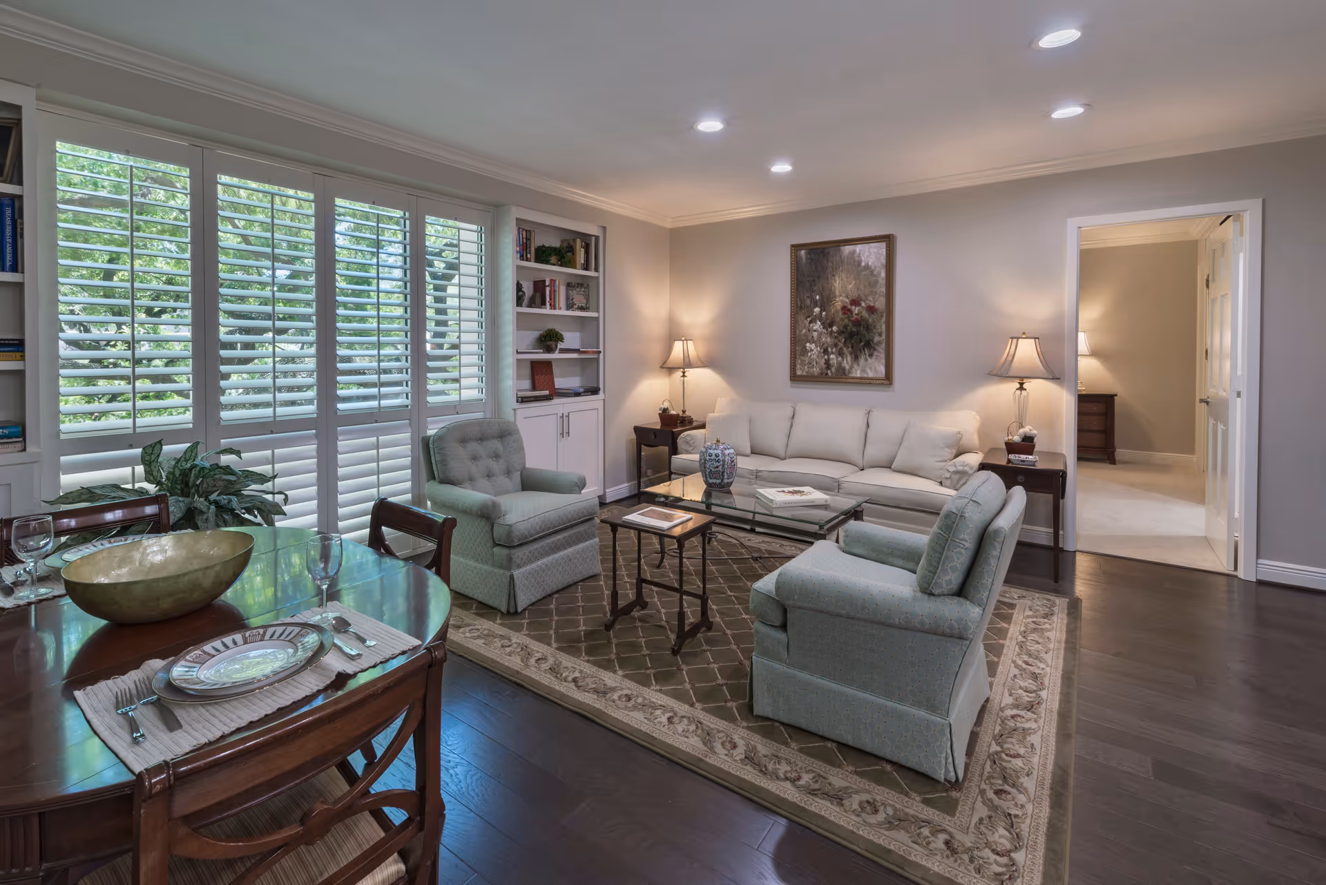 Bright living room with a white sofa, two upholstered armchairs, glass coffee table, rug, built-in shelves, and a dining table by shuttered windows.