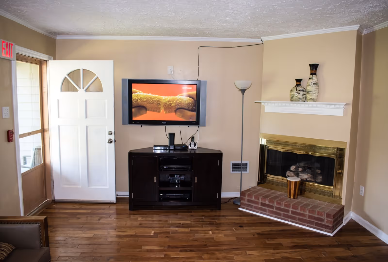 Cozy living room with a wall-mounted TV above a media cabinet, a white front door to the left, and a fireplace with decorative vases on the right.