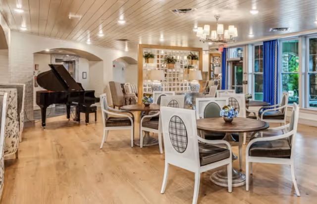 A spacious and well-lit common area with wooden floors, round tables surrounded by white chairs with dark cushions, a grand piano in the corner, large windows with blue curtains, and decorative lamps and plants.