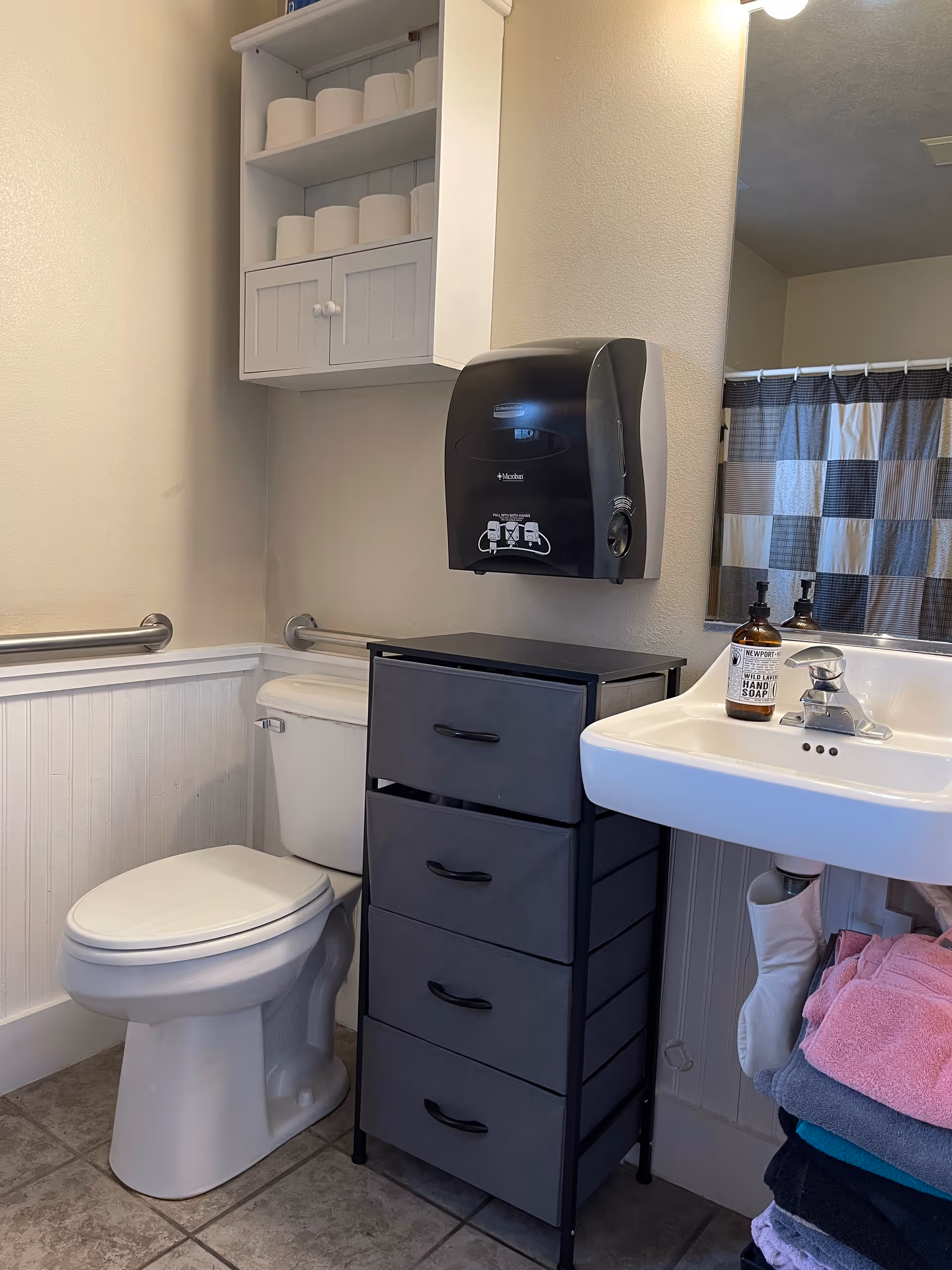 A clean bathroom featuring a white toilet with a grab bar on the wall, a white wall-mounted cabinet stocked with toilet paper rolls, a black paper towel dispenser, a black four-drawer storage unit, and a white sink with a bottle of hand soap. A mirror above the sink reflects a checkered shower curtain.